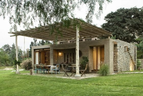 Fachada de casa rústica con adobe y piedra, tiene un techo de caña y tronco en patio exterior